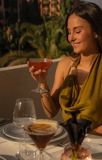 Femme assise sur la terasse de la Villa Chakamani, souriante, au soleil, tenant un cocktail signature à la main